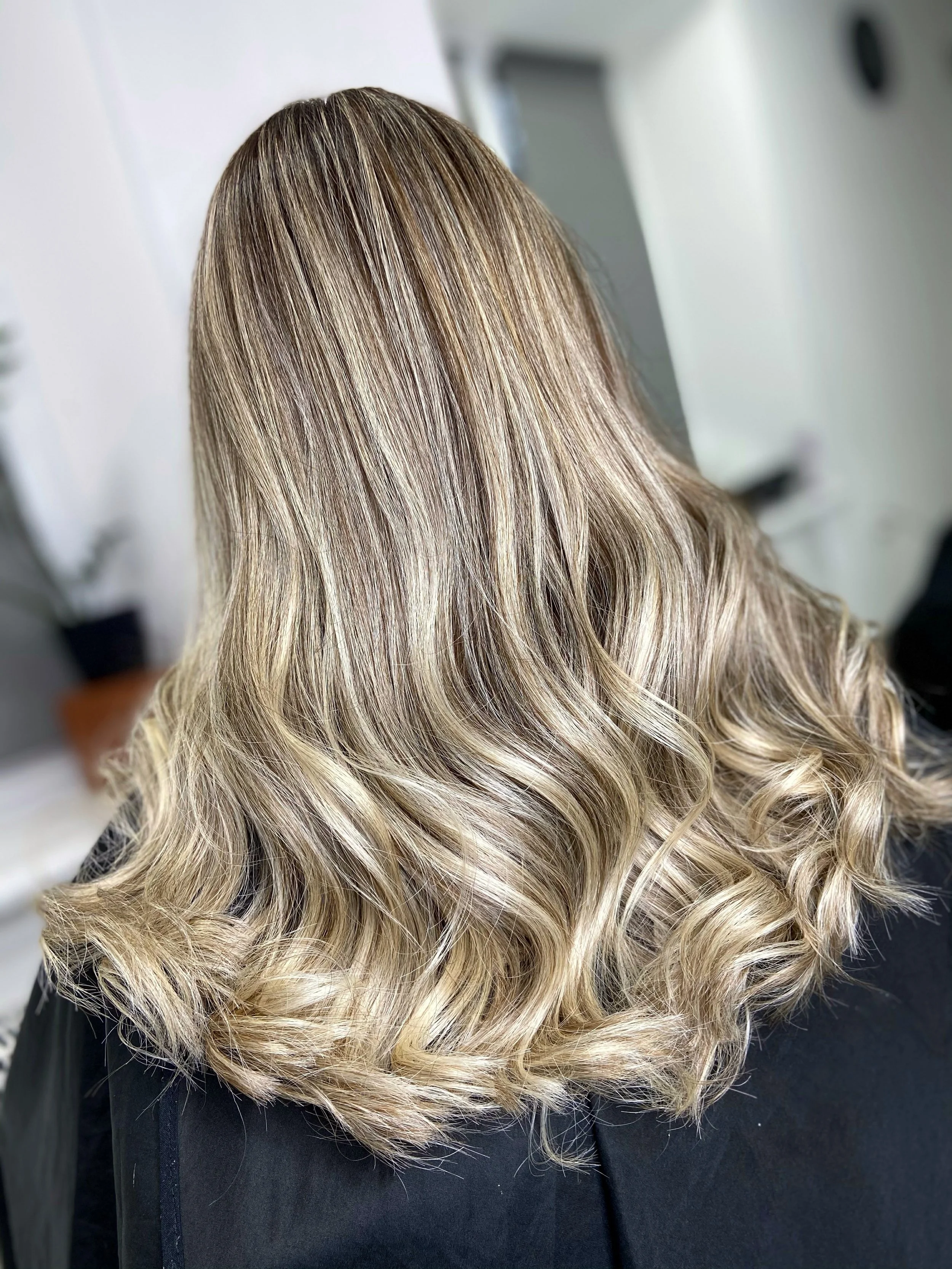 Back view of a woman with shoulder-length, wavy blonde hair in a salon.