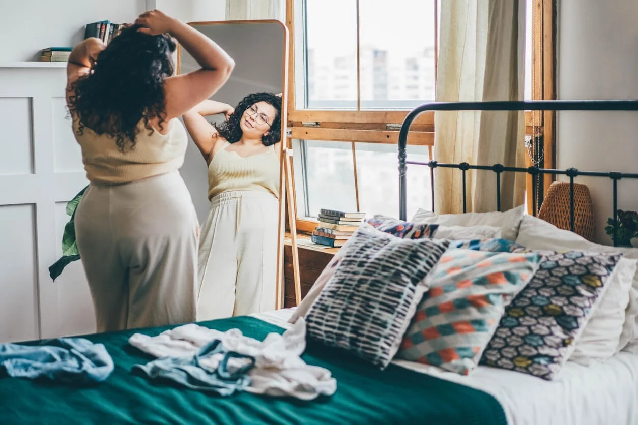 A woman in a cream top and wide-leg pants fixes her hair in a bedroom mirror beside a bed with patterned pillows and clothes laid out, getting ready for a date