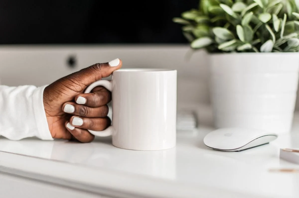 A hand holding a white mug at a clean desk beside a small plant, a calm start to the day.Boundary script graphic: Acknowledge, set the Boundary, then offer a Creative Yes when a parent criticizes your choices.