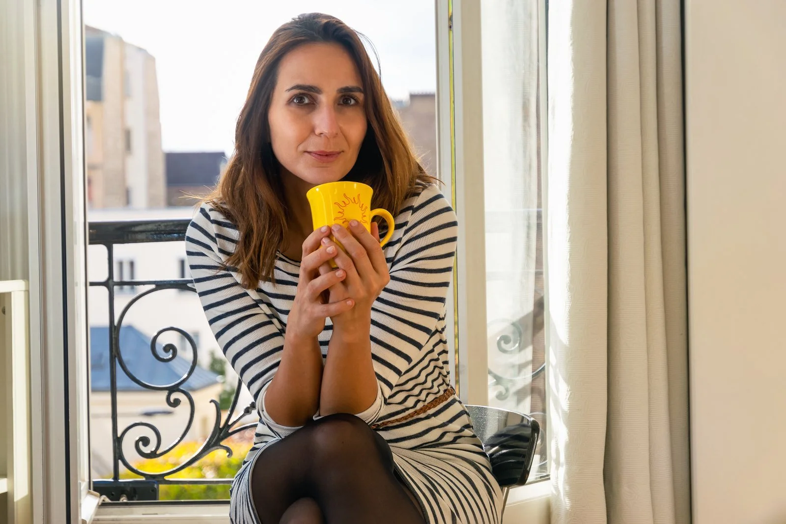 A smiling woman holding a mug by a sunny window, enjoying a calm morning routine that supports self-confidence.