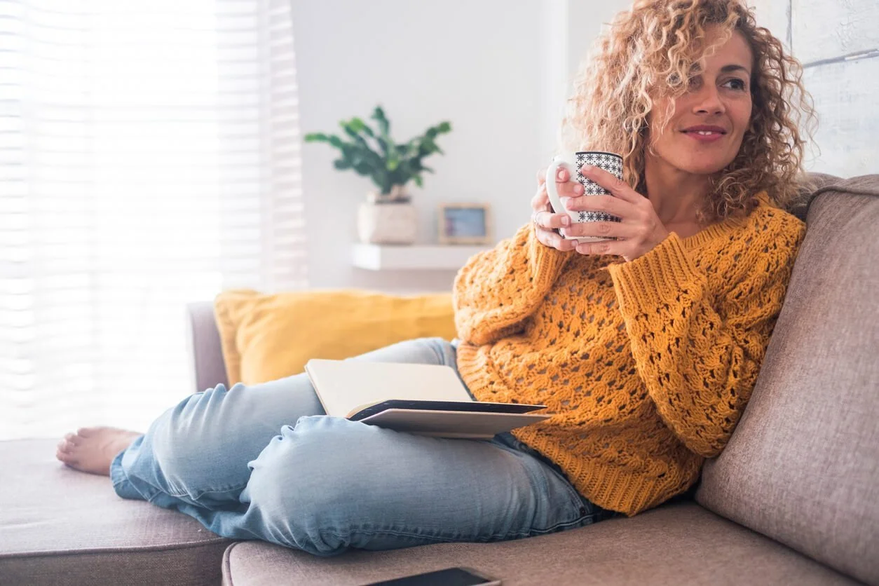 A woman with curly blond hair in an orange knit sweater and jeans relaxes on a sofa holding a mug, an open book on her lap in a bright living room