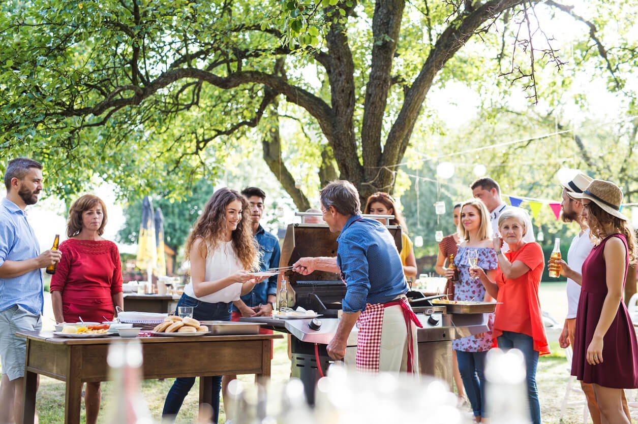 Adults gather around a backyard barbecue under tall trees on a summer day, a man in a blue shirt at the grill while others chat and hold drinks