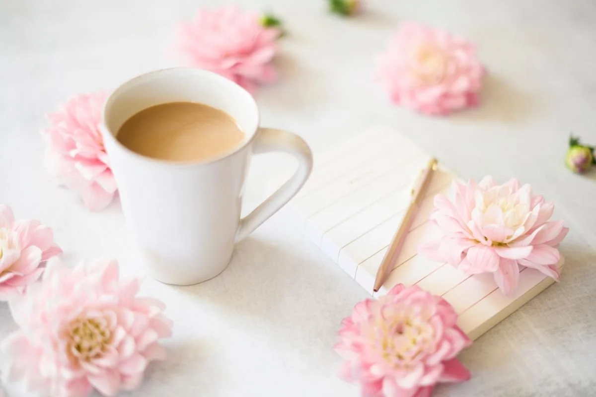 A cup of tea beside a notebook and pink flowers on a white table, a quiet moment of comfort.