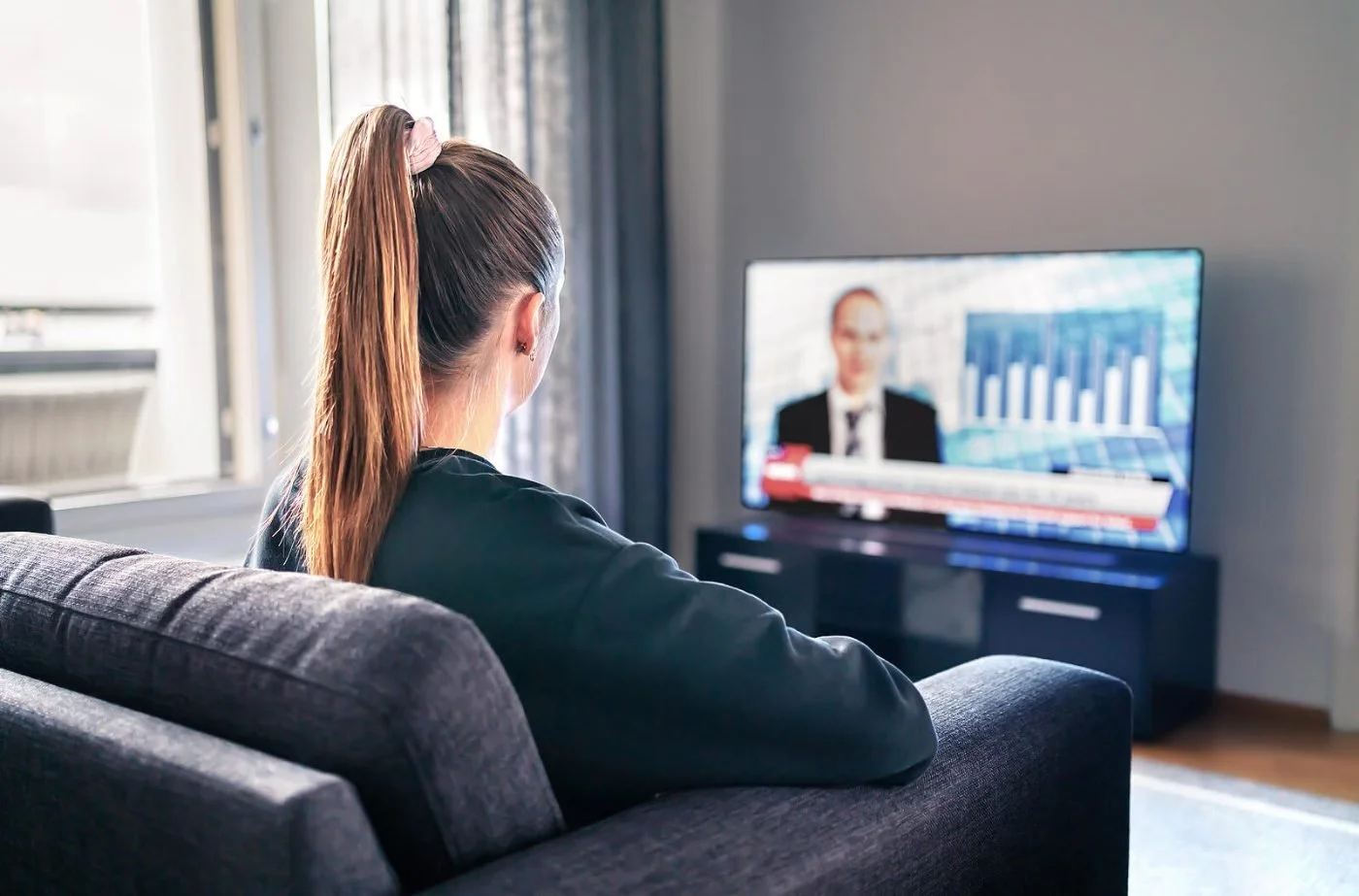 A woman with a ponytail sitting on a couch watching a news broadcast on television.