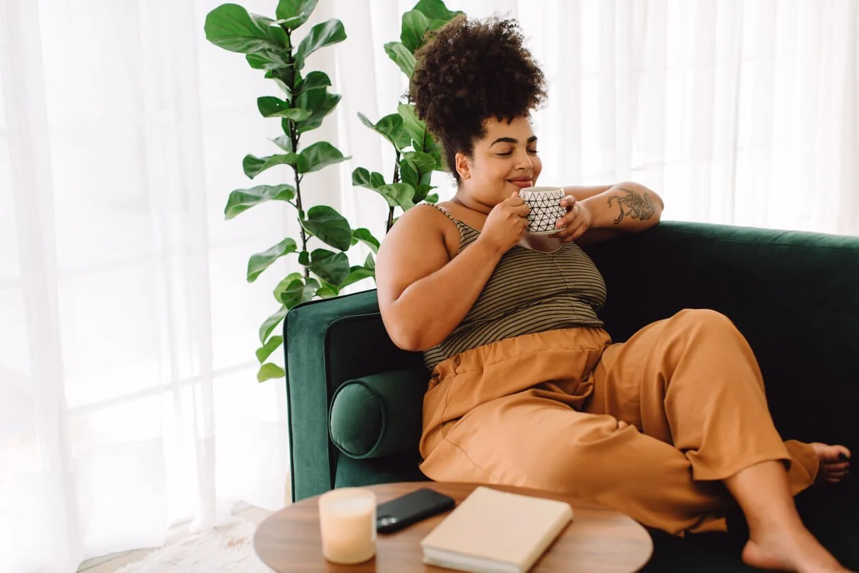 A smiling woman with her hair in a curly bun sips from a mug in a deep green armchair beside a large leafy plant, a candle, a phone, and a book on the table in front of her