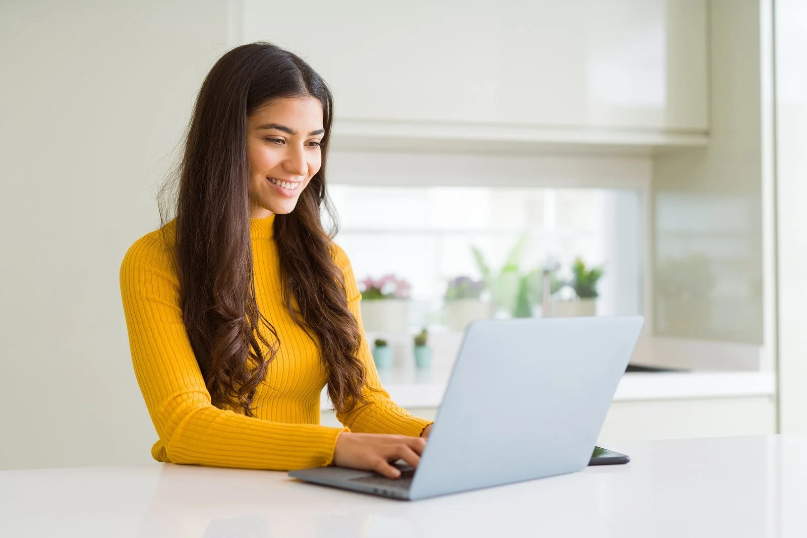 A smiling woman in a yellow sweater sitting at a kitchen counter with her laptop, ready for an online therapy session.