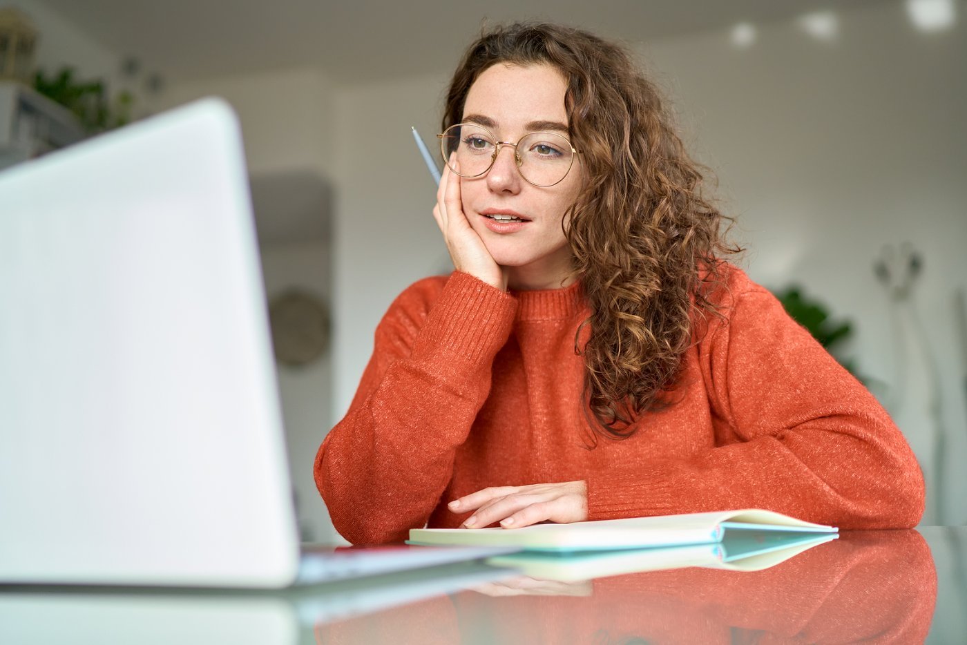 A woman in glasses and an orange sweater rests her chin on her hand at her laptop, looking lost in thought as she works.