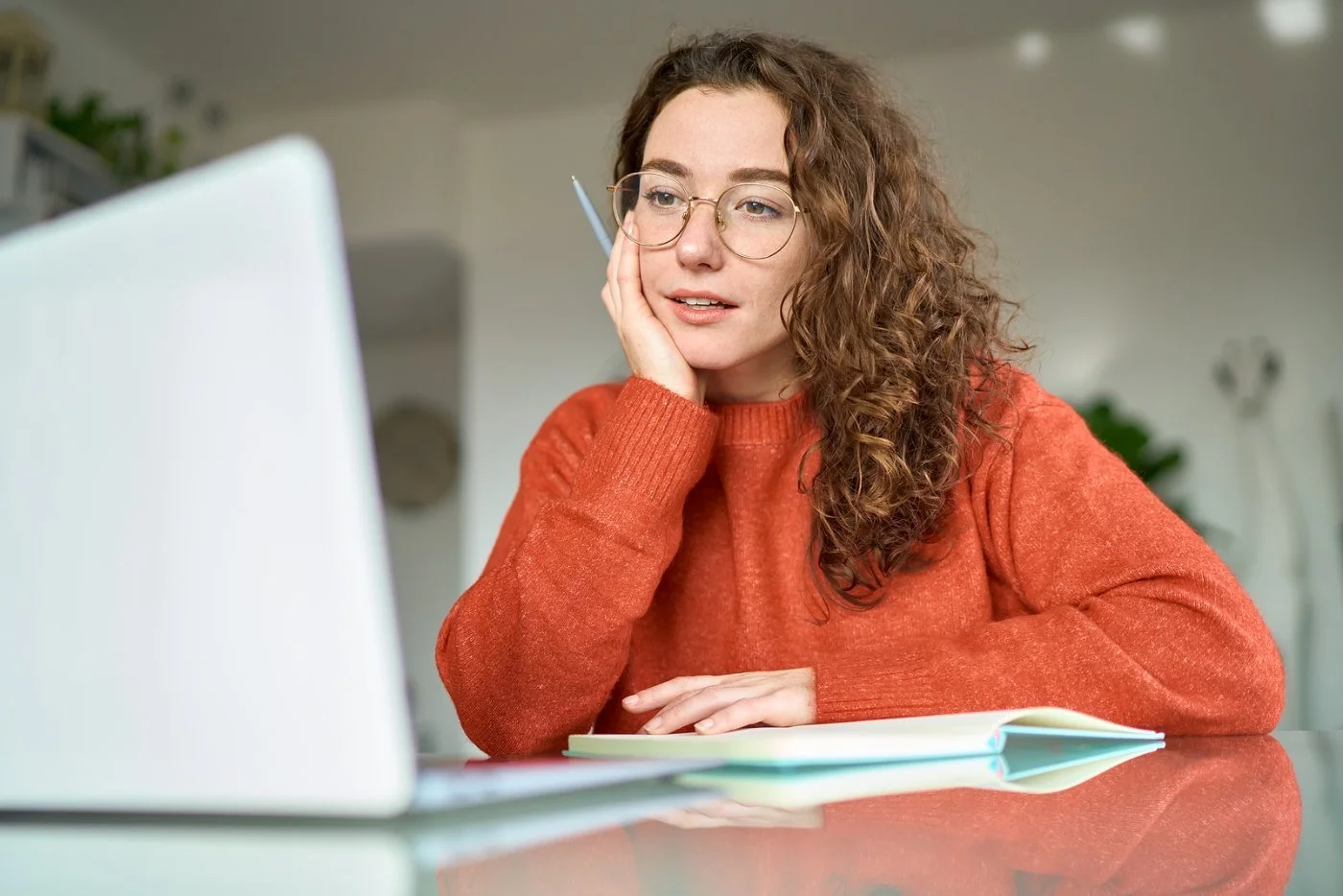 A young woman with curly hair and glasses in an orange sweater rests her cheek on her hand beside a laptop, looking stuck on a task