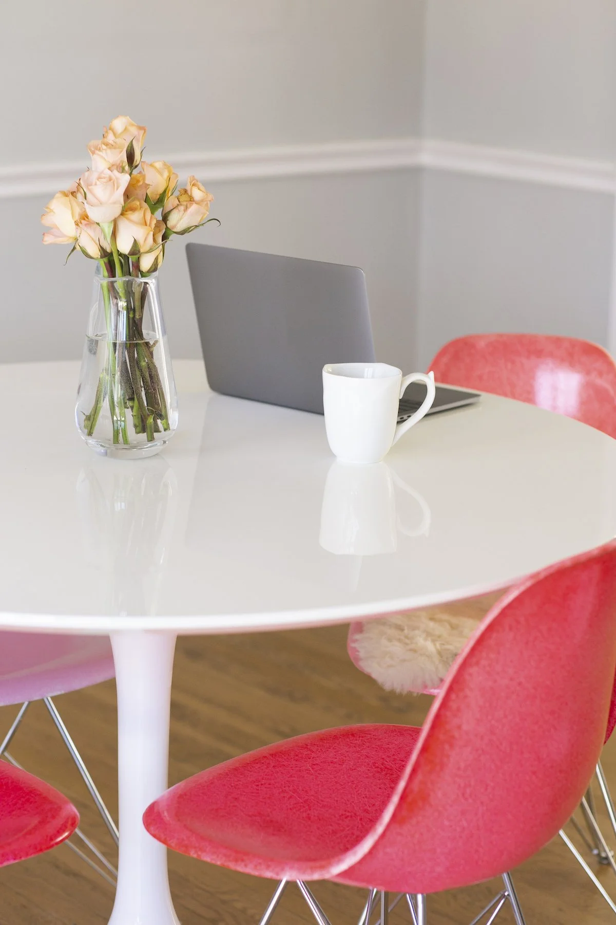 A white table with pink chairs, a laptop, flowers, and a coffee cup, set up for an online therapy session.