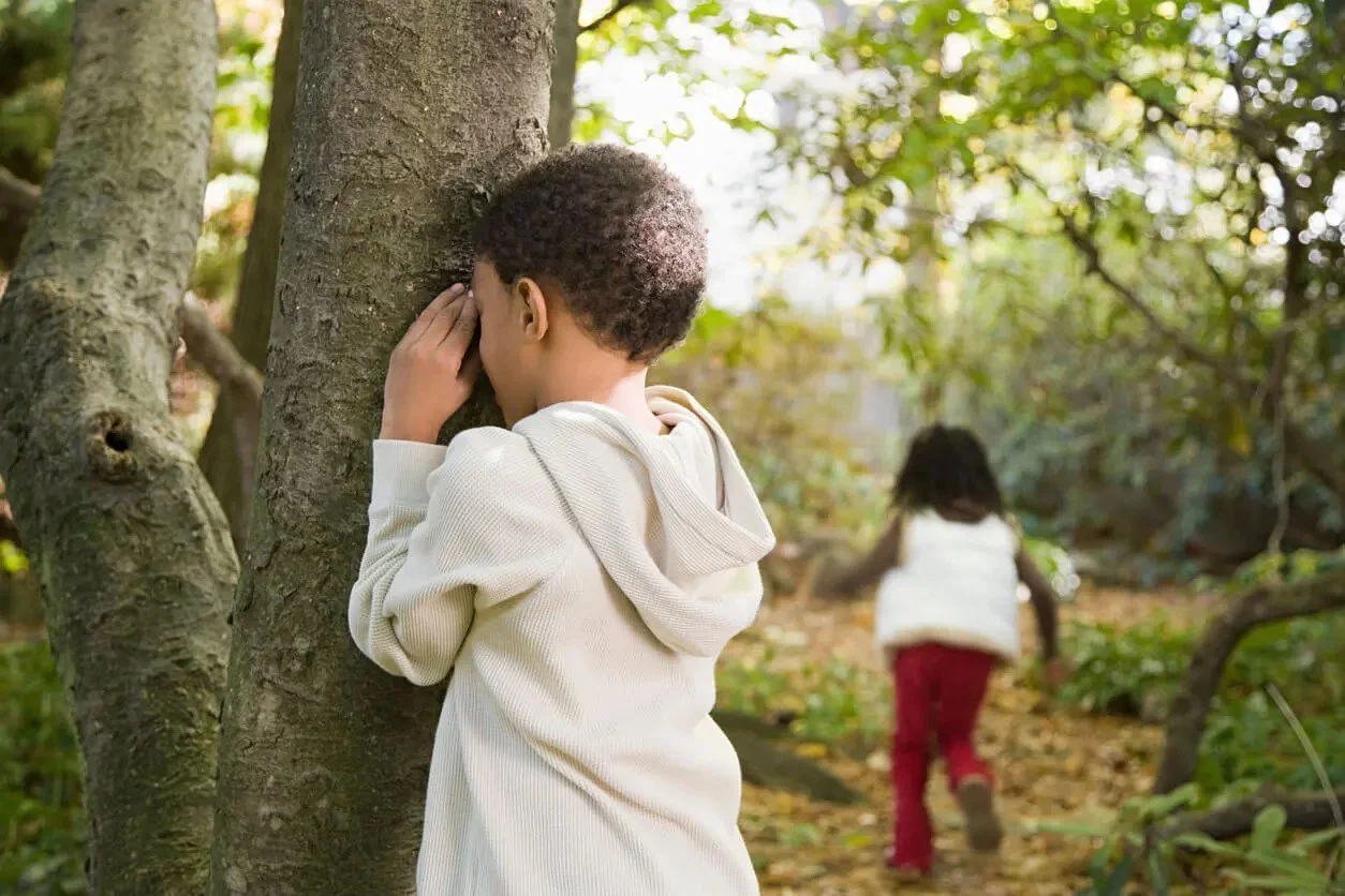 A boy counting with his hands over his eyes beside a tree while a girl runs off to hide, a game of hide and seek.