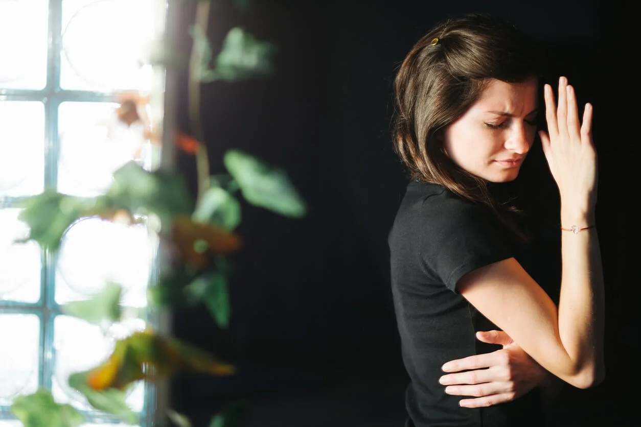 A woman in a black top standing by a bright window with her hand raised to shield her face from the light, coping with light sensitivity.
