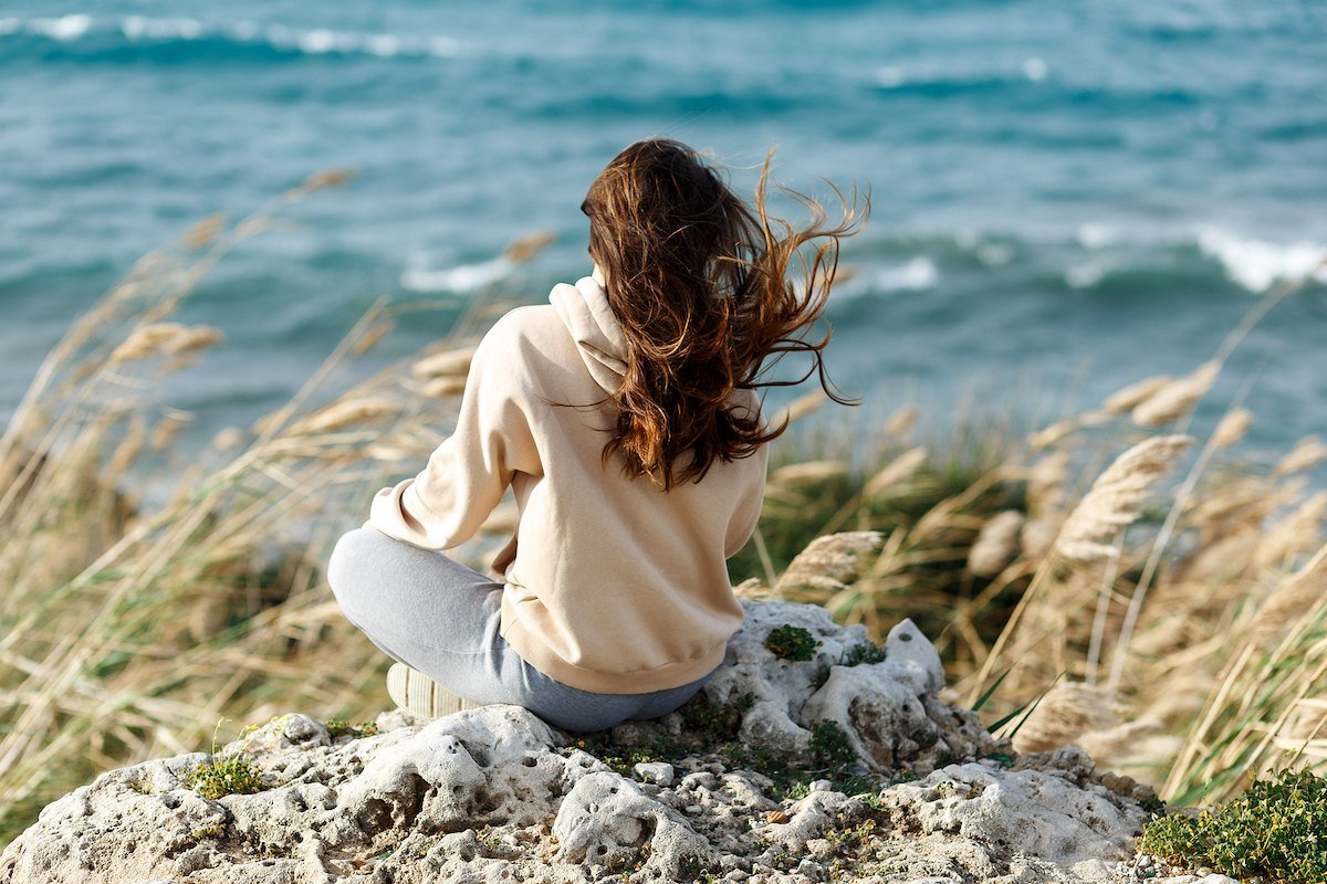 A woman in a cream sweater sitting on a rocky seaside looking out at the waves, finding her footing after divorce.