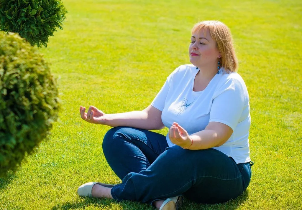 A woman sitting cross-legged on a sunny lawn with her eyes closed, taking a quiet moment to recharge.