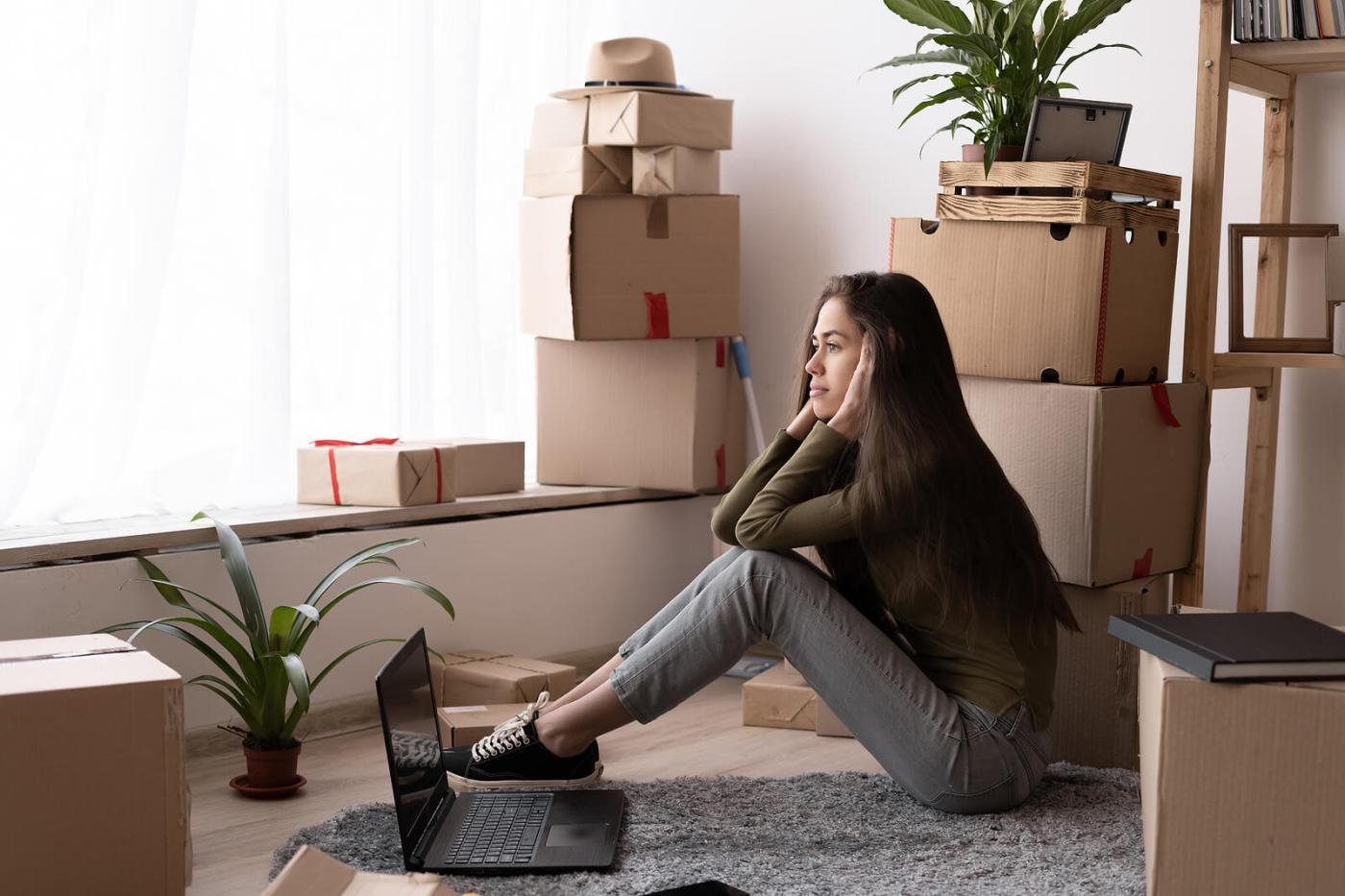 A woman sitting on the floor of a room stacked with moving boxes, looking tired in the middle of a move.