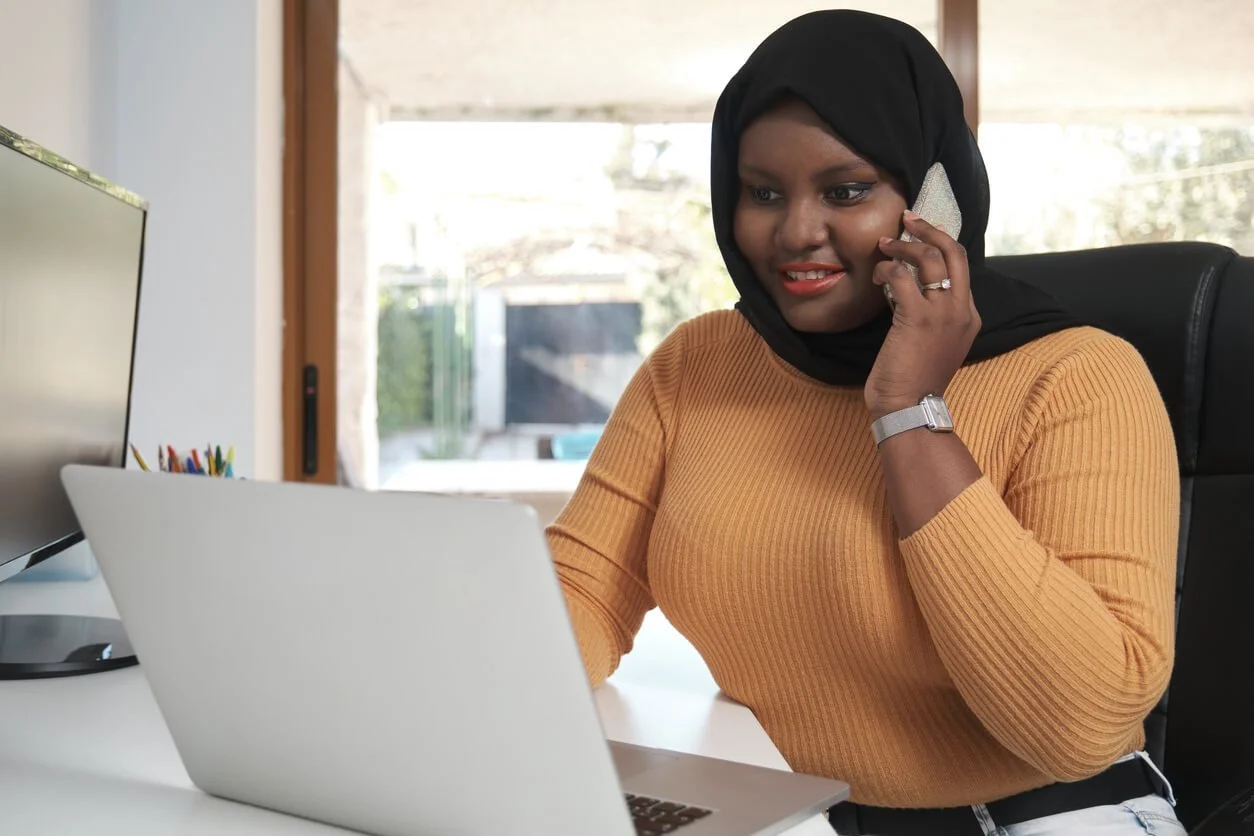 A smiling woman in a black hijab and orange knit sweater talks on the phone while looking at her laptop at a desk in a bright office