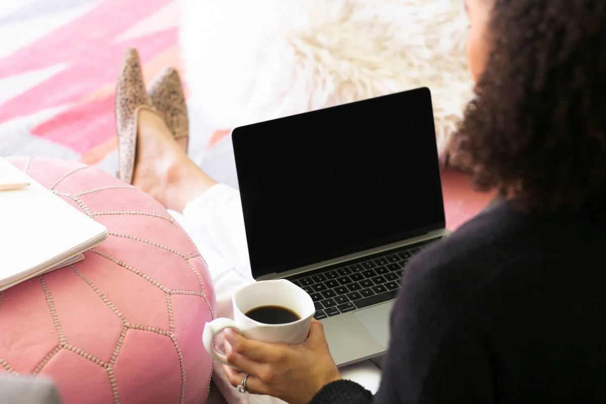 A woman with her feet up working on a laptop and holding a cup of coffee beside a pink pouf, a relaxed picture of work-life balance