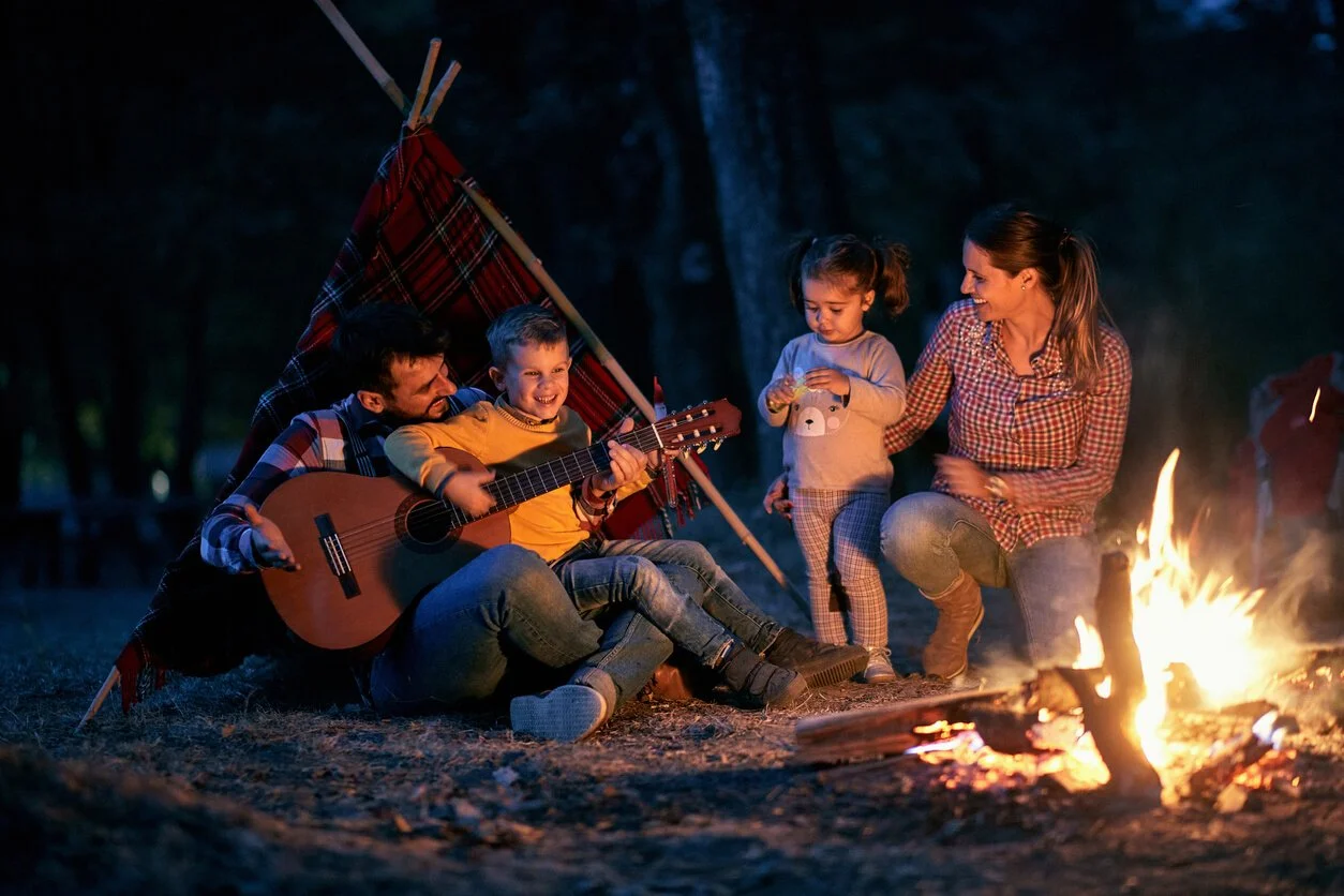 A family sitting by a campfire while one parent plays the guitar, a cozy scene from a camping trip.