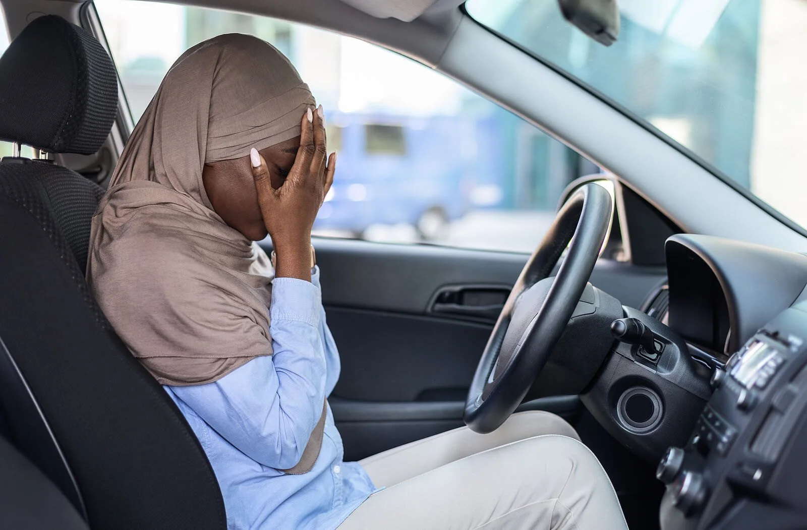 A woman in a headscarf sitting in her parked car with her hands over her face, overwhelmed by depression.