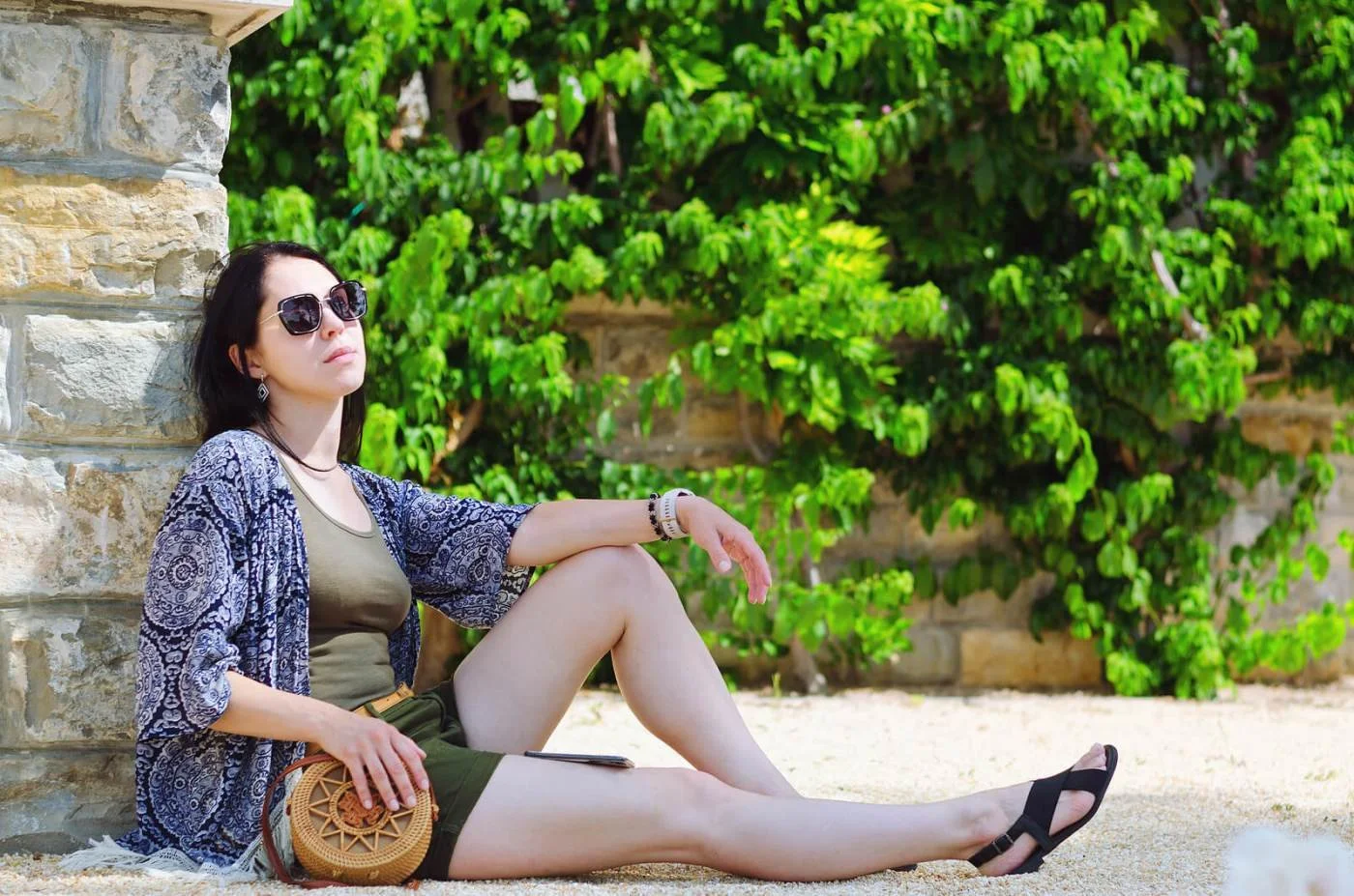 A woman in sunglasses sitting outside in front of a stone wall and green foliage, taking a break from the sun.