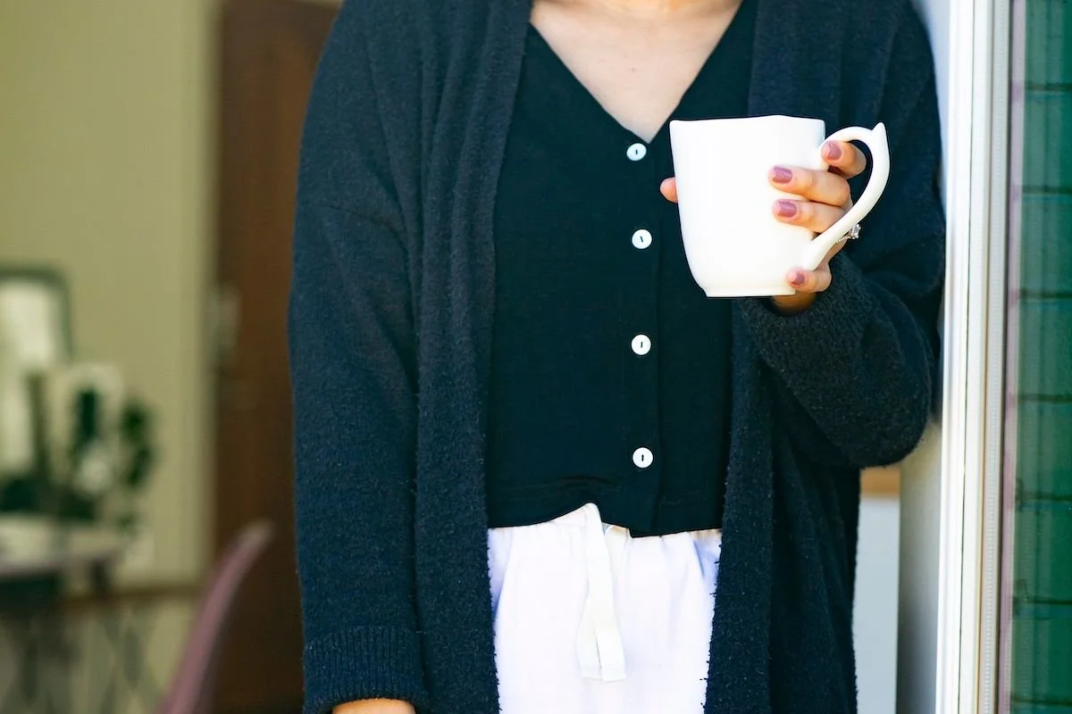 A woman in a dark cardigan holding a white mug in a doorway, taking a quiet moment of self-care.