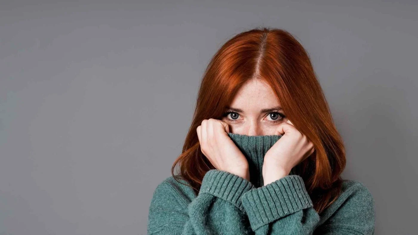 A woman with red hair pulling her teal sweater up over her nose and looking shyly at the camera.