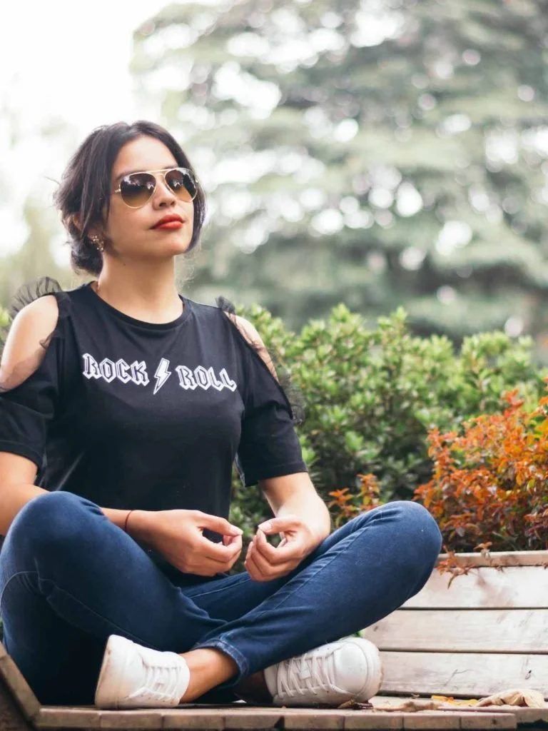 A woman in a rock and roll t-shirt sitting outdoors with her hands in her lap, taking a calm moment among the plants.