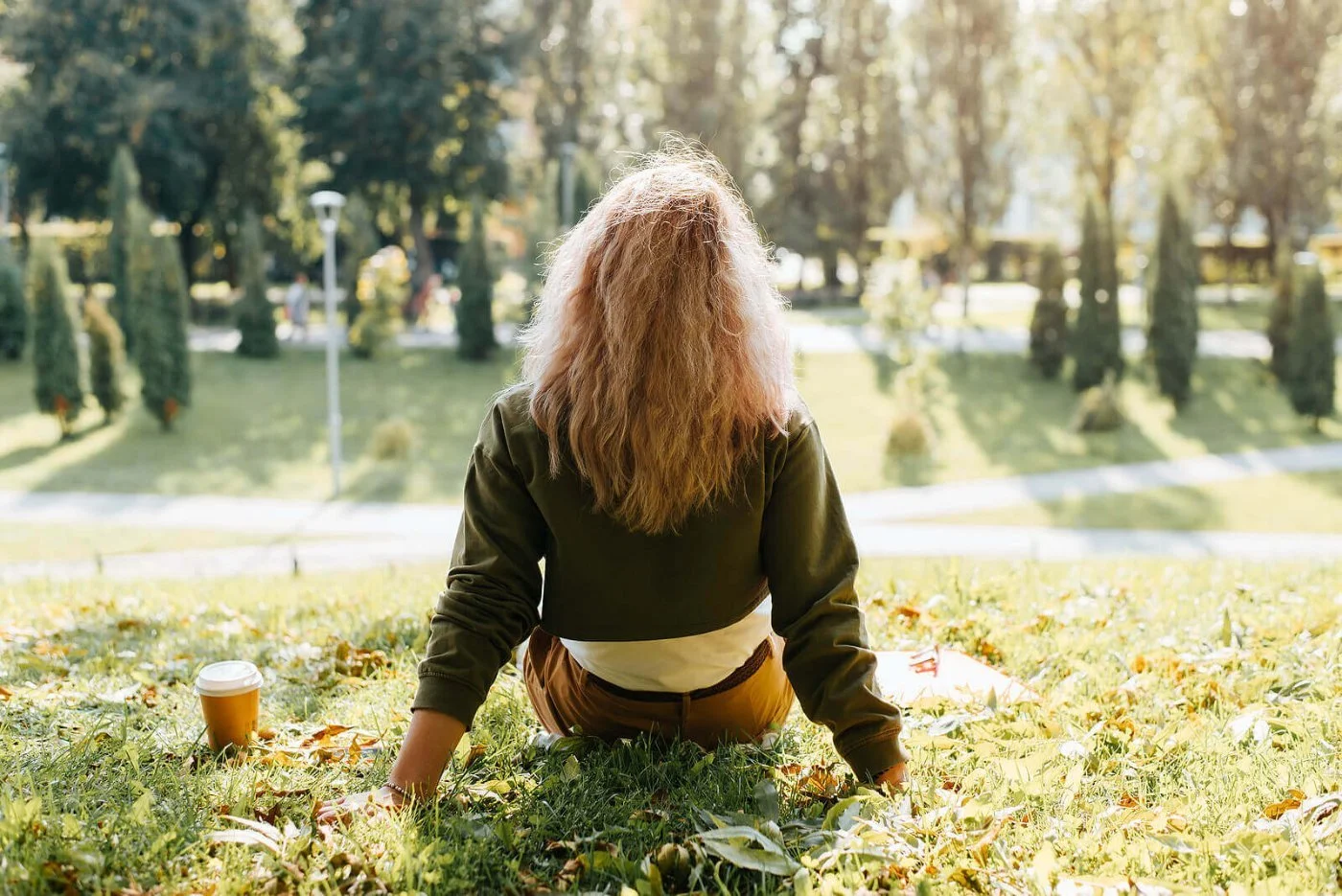 A woman with wavy hair seen from behind sits on a park lawn scattered with autumn leaves, a takeaway coffee cup in the grass beside her
