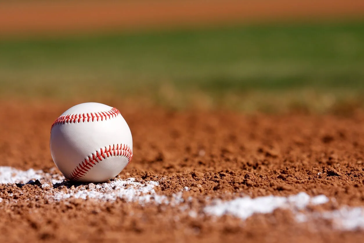 A white baseball with red stitching rests on the dirt beside a chalk line on a baseball field, a nod to the curveballs life throws at highly sensitive women