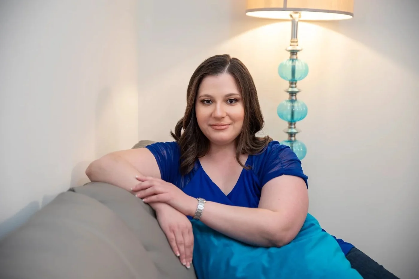 Enid DeJesus, a licensed mental health counselor, in a blue top seated on a gray sofa beside a lamp with teal glass details