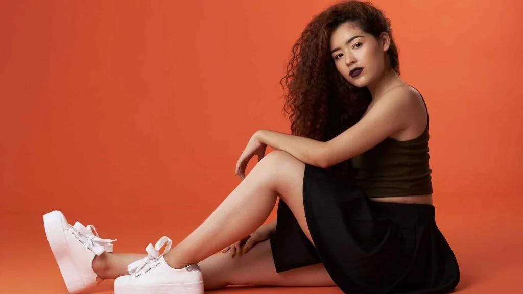 A woman with long curly hair sitting in white sneakers against an orange backdrop, relaxed and expressive.