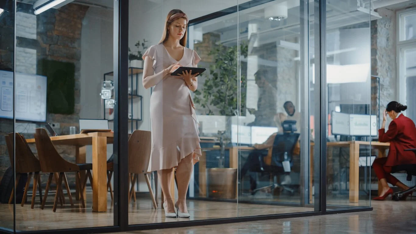 A woman in a pale pink dress stands holding a tablet inside a glass-walled open office while colleagues work at desks behind her