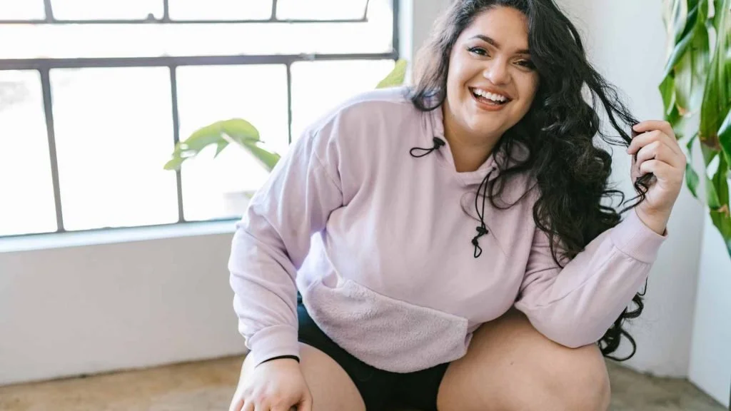 A smiling woman with long dark hair in a pink hoodie crouching in a bright room, at ease with herself.