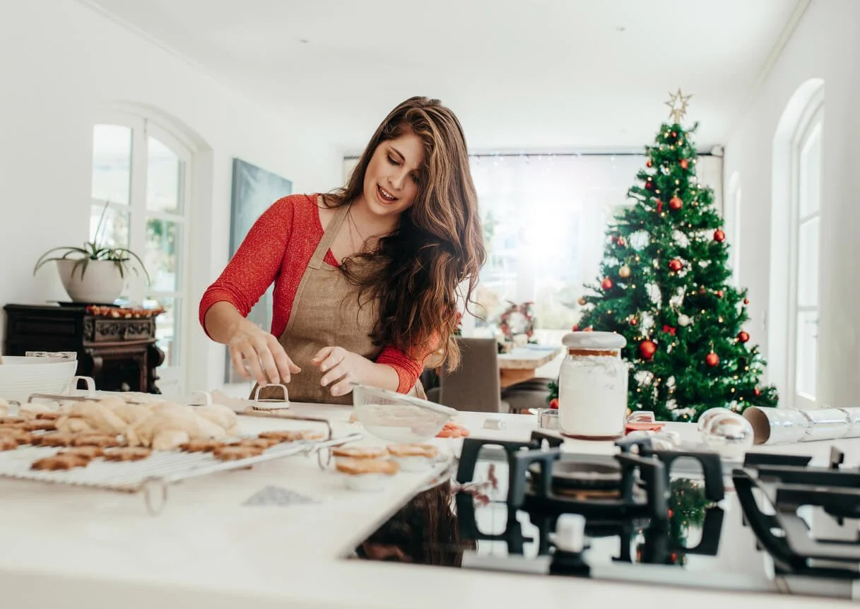 A woman in a red cardigan and apron prepares cookies at a bright kitchen counter with trays of baked cookies and a decorated Christmas tree behind her
