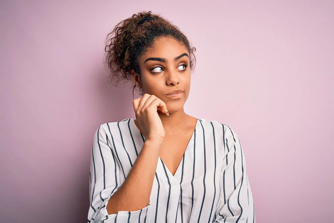 A woman in a striped blouse against a pink wall with her hand on her chin, looking thoughtful as she weighs the cost and value of therapy