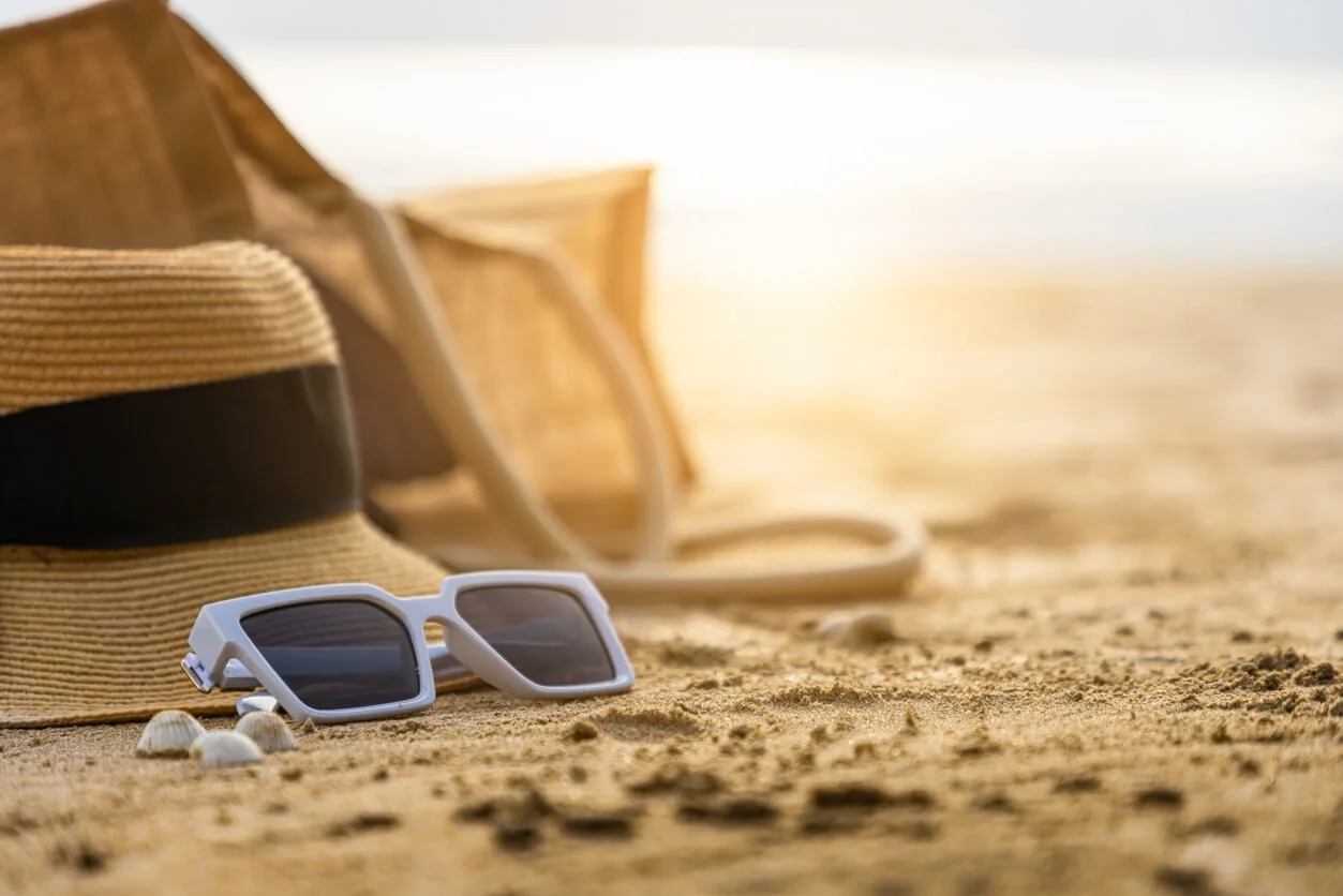 A straw hat and white sunglasses resting on sunlit sand at the beach, the kind of gear that eases bright days.