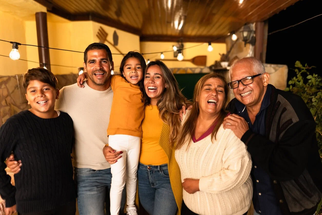 A laughing multi-generational family of two children, two parents, and two grandparents hugging close together on an evening patio