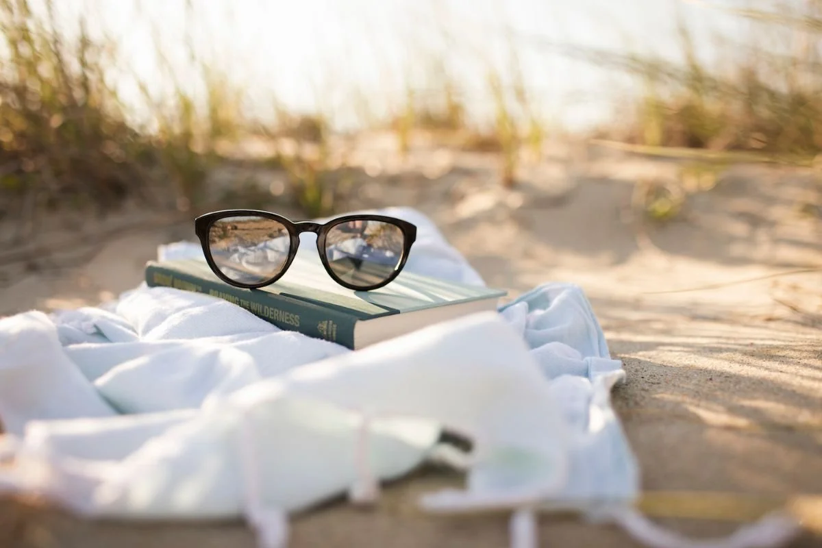 A pair of glasses resting on a book and a blanket in the beach dunes, a reminder to step away and rest.