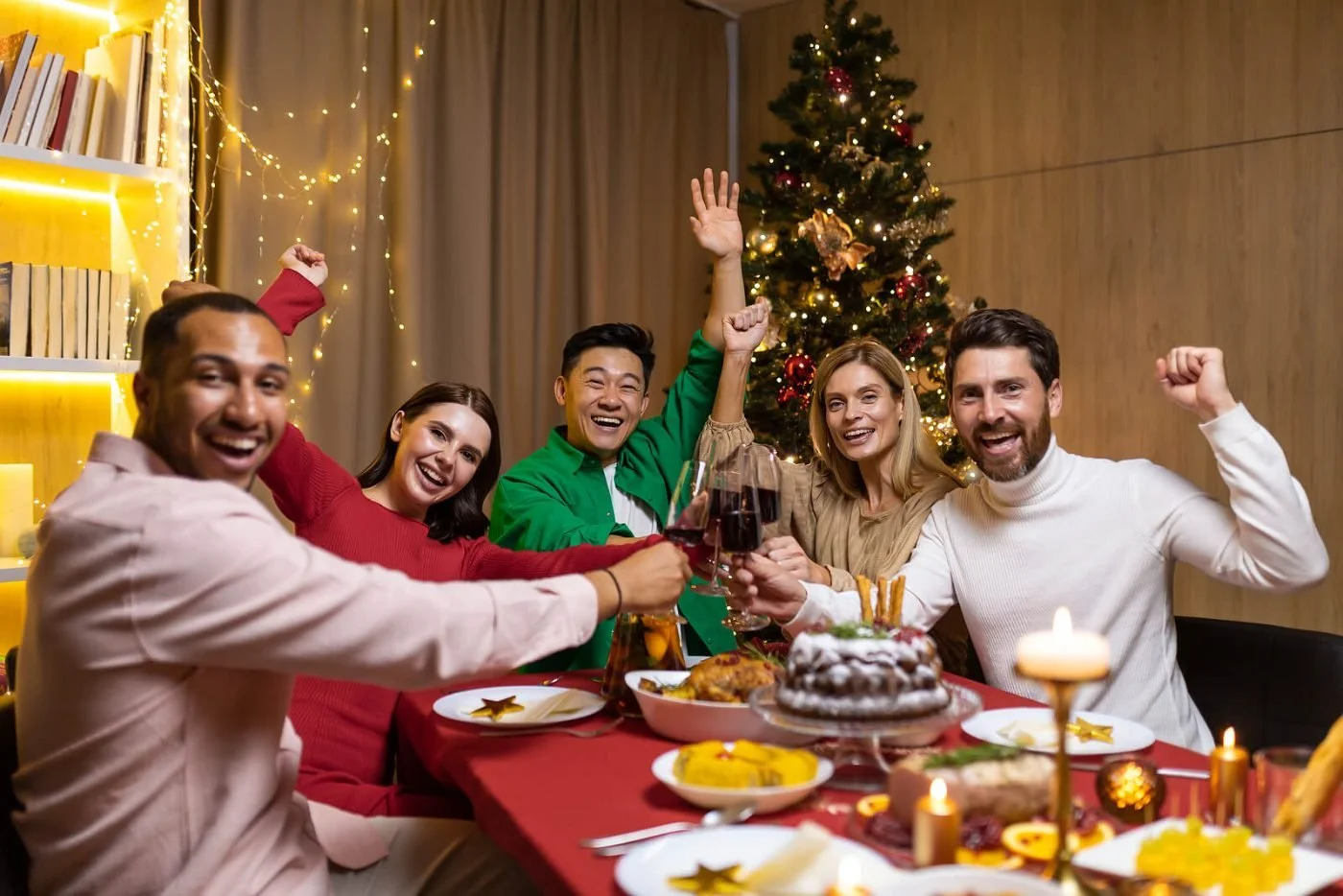Friends cheering around a festive holiday dinner table with a decorated tree in the background.