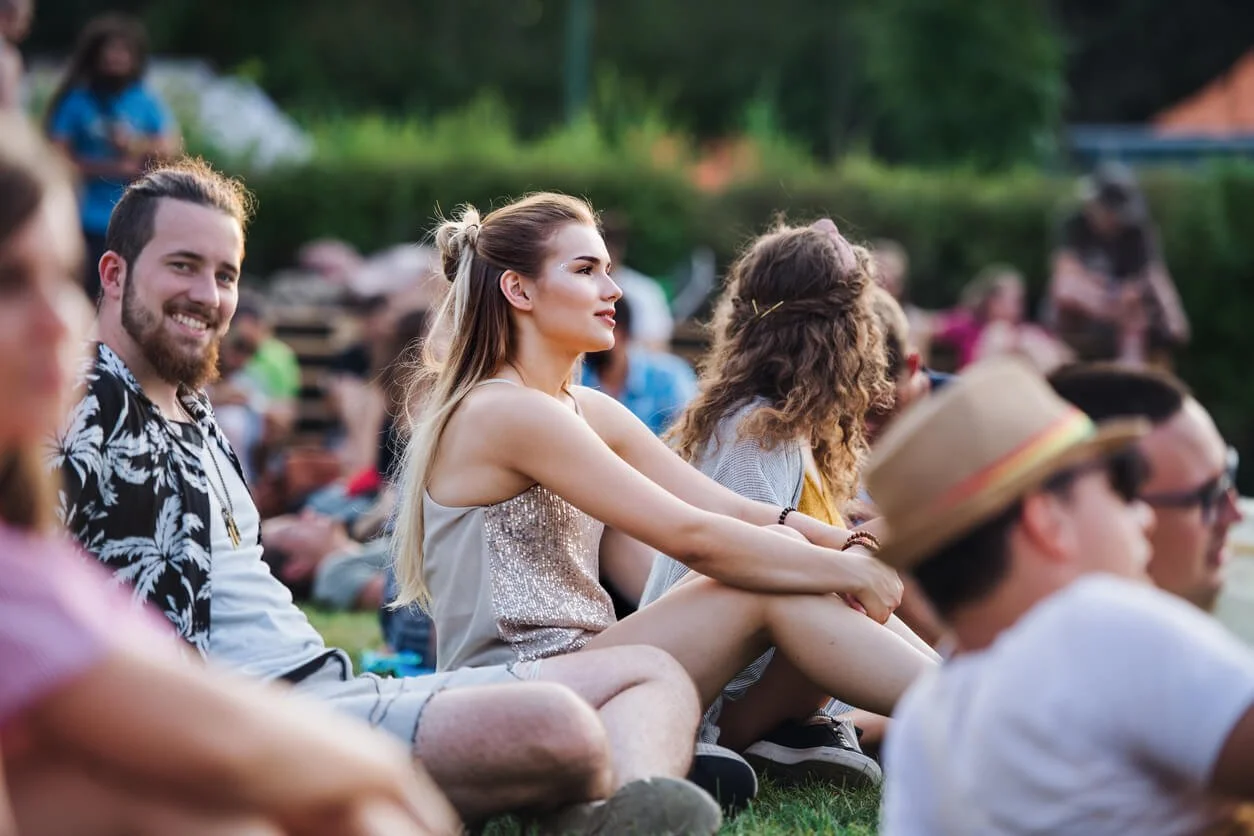 A woman sitting on the lawn among a relaxed crowd and smiling toward a concert stage, enjoying the music from a comfortable spot.