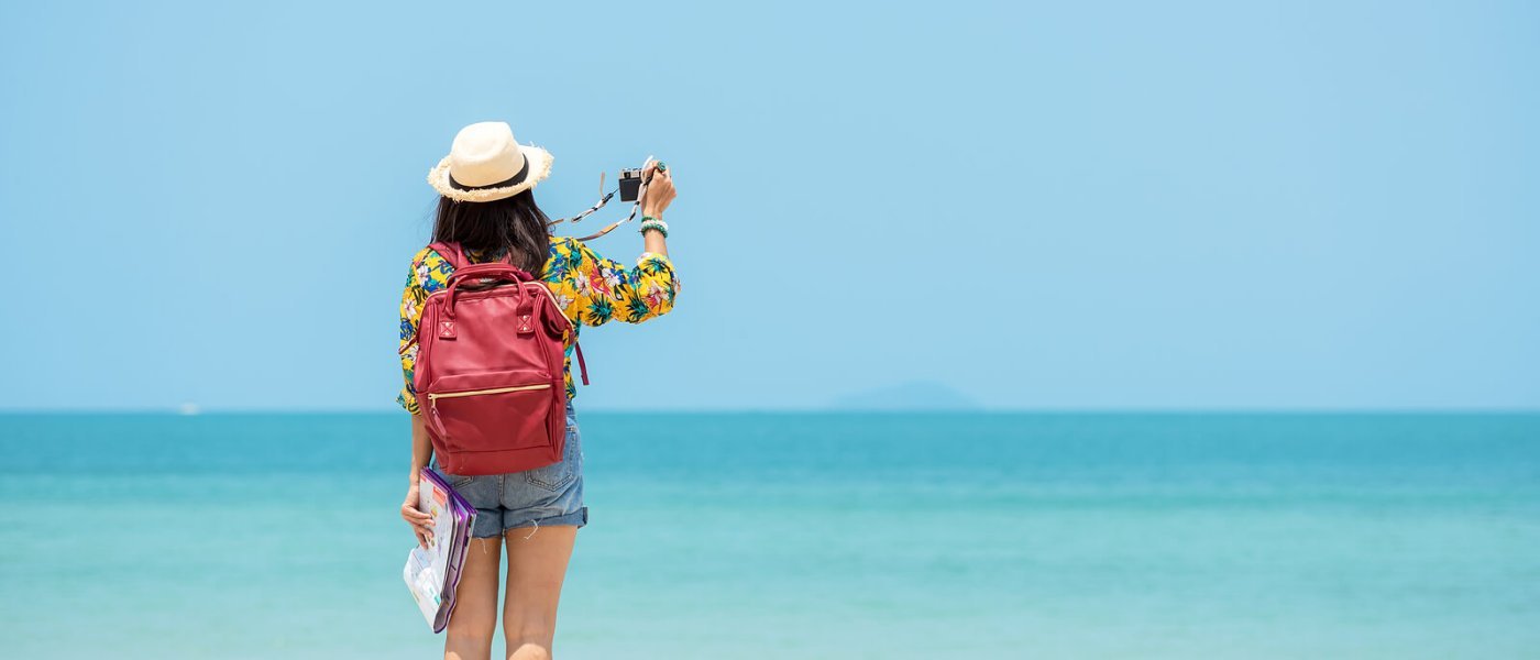 A woman in a sun hat and backpack standing by the ocean and taking a photo on a solo trip, traveling at her own pace.