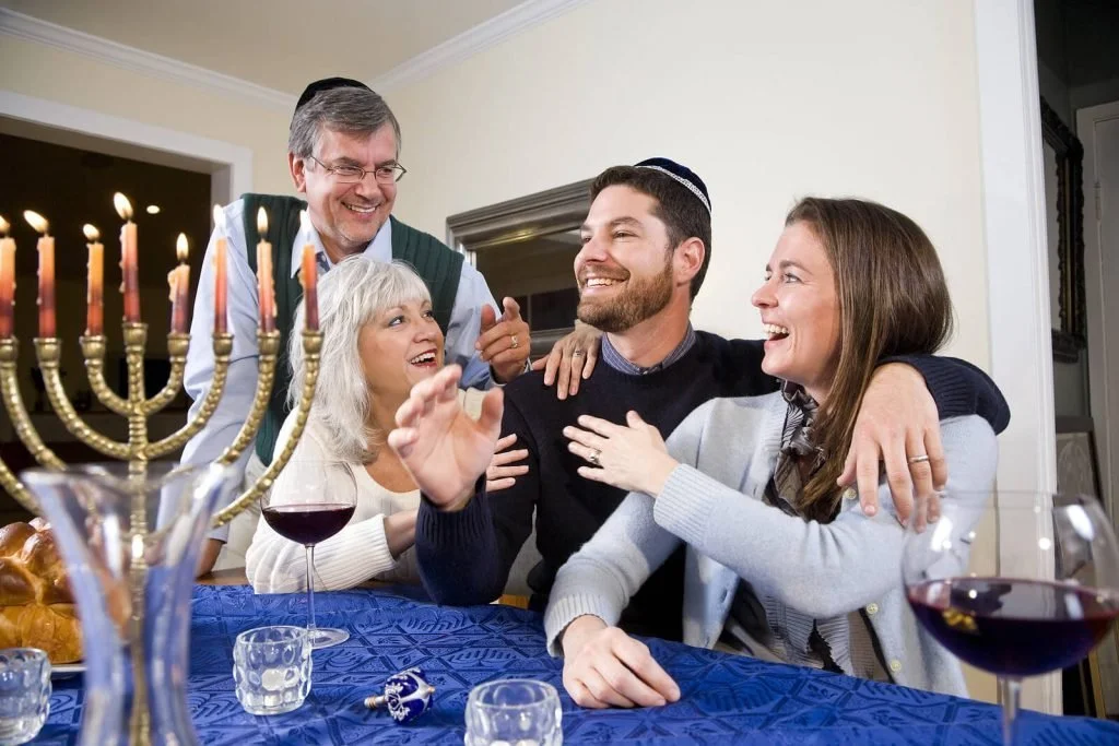 A family gathered around a table with a menorah and wine glasses, laughing together during a Hanukkah celebration.