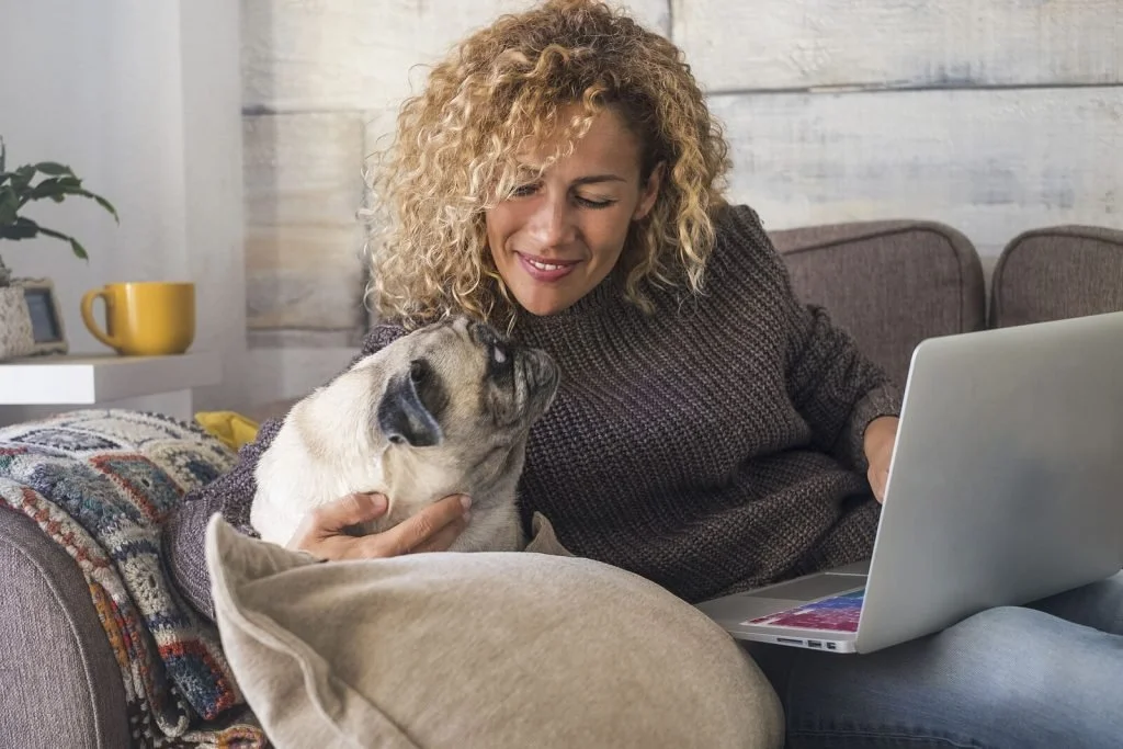 A woman with curly hair working on a laptop on her couch while a pug rests beside her, an energy-protecting way to work from home.