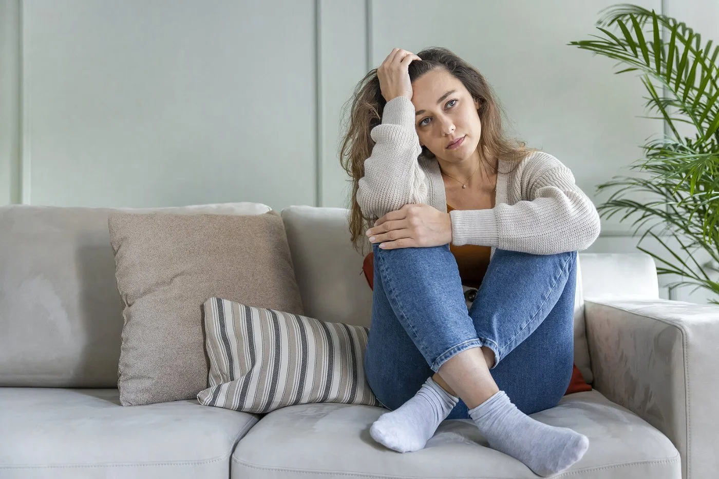 A woman sits on a gray sofa with her knees pulled up and one hand in her hair, looking tired and weighed down by grief