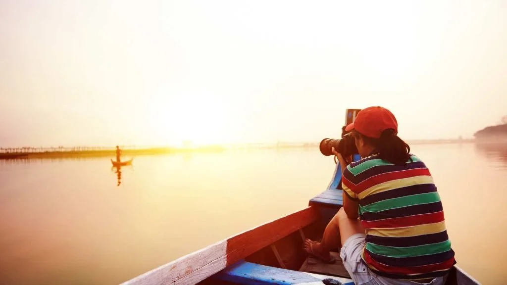 A woman in a colorful striped shirt and red cap taking a photo from a canoe at sunset, enjoying travel at a calm pace.