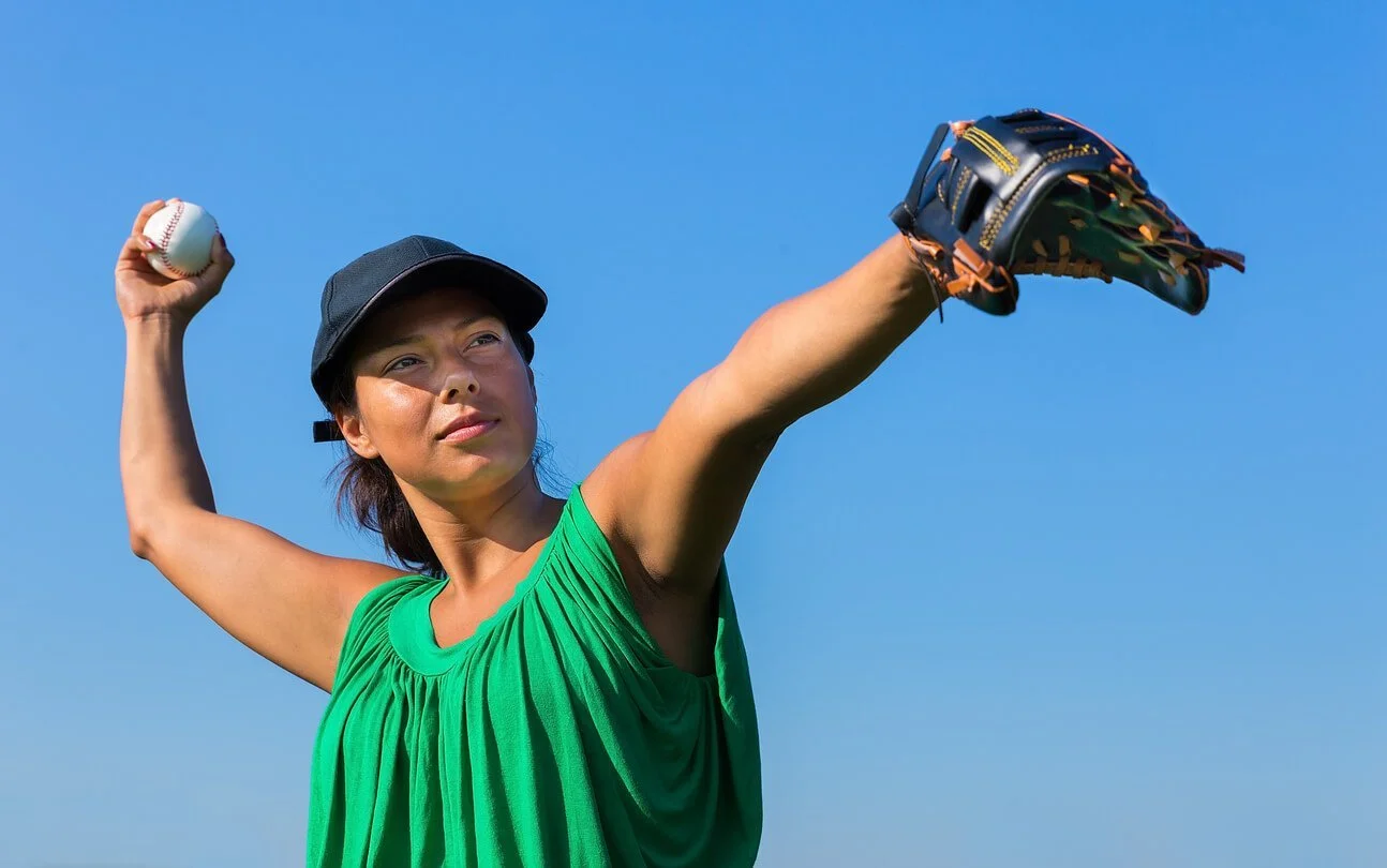 A woman in a green sleeveless top and dark cap against a clear blue sky holds a baseball in one hand and stretches her gloved hand out to the side