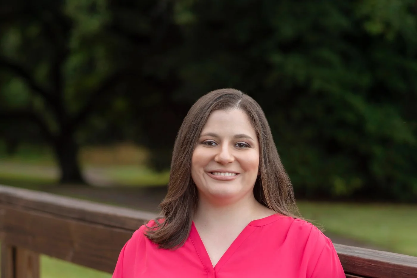 Enid DeJesus, a licensed mental health counselor, smiling in a bright pink top beside a wooden fence with green trees behind her