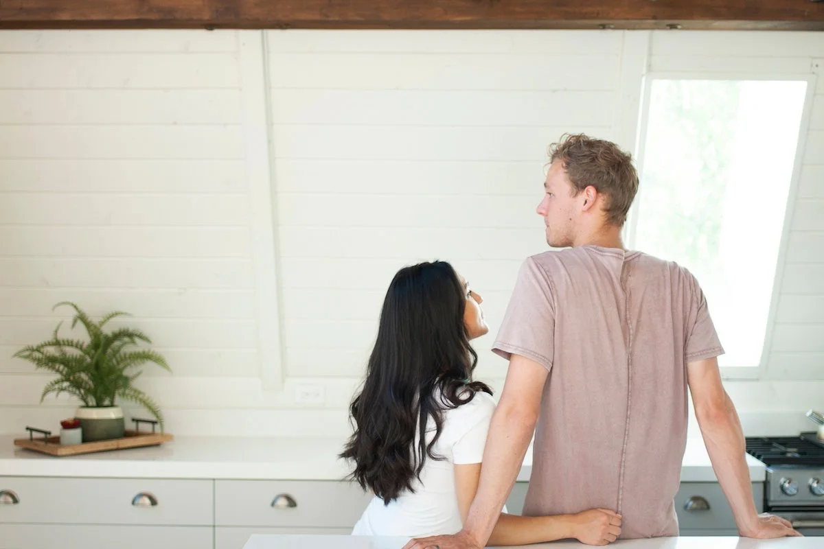 A couple seen from behind leaning on a kitchen counter, the woman turned toward the man as they talk, a relaxed moment where an honest conversation can happen