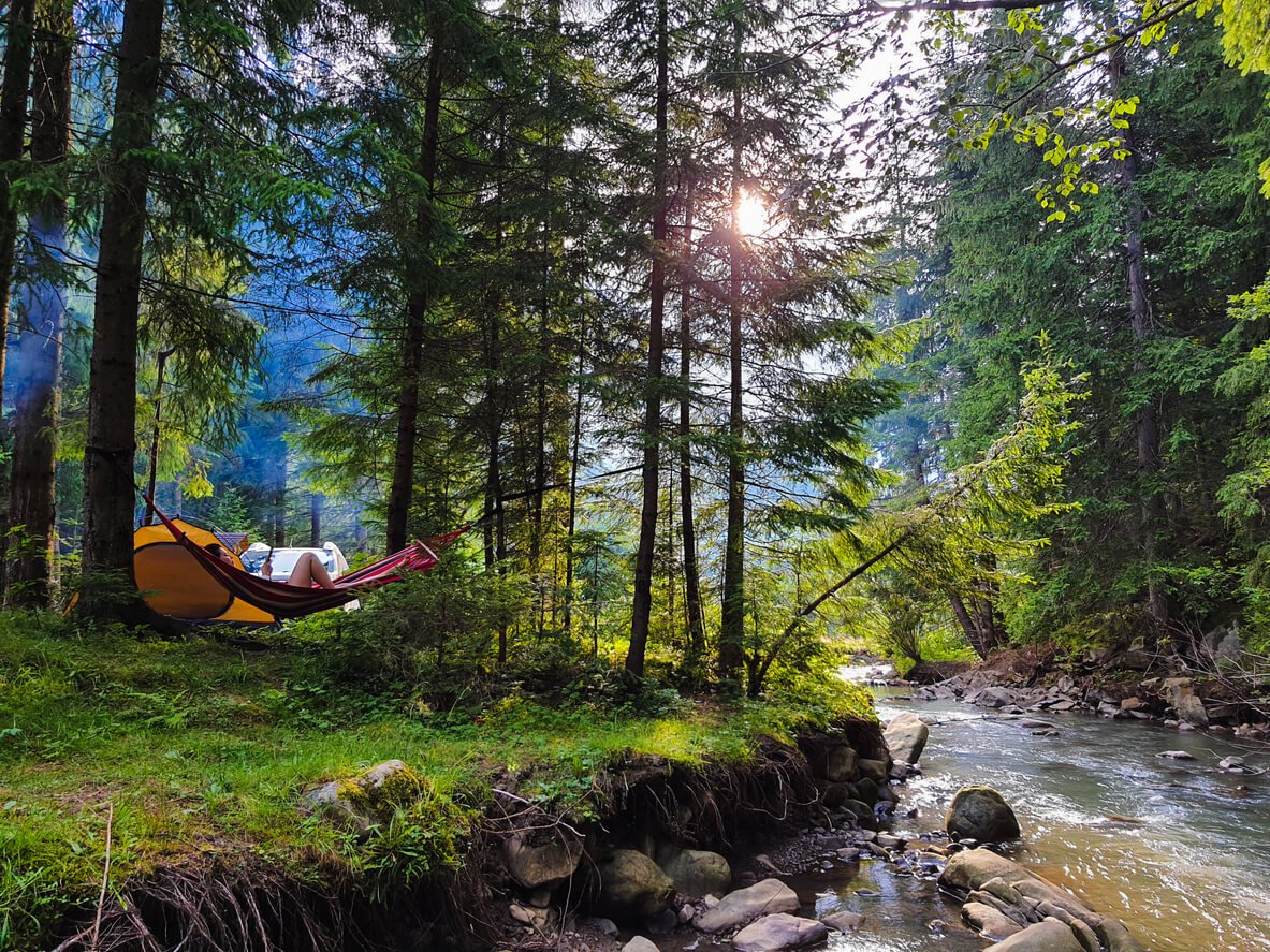 A woman relaxing in a hammock by a creek in the woods, enjoying a camping trip as an introvert.