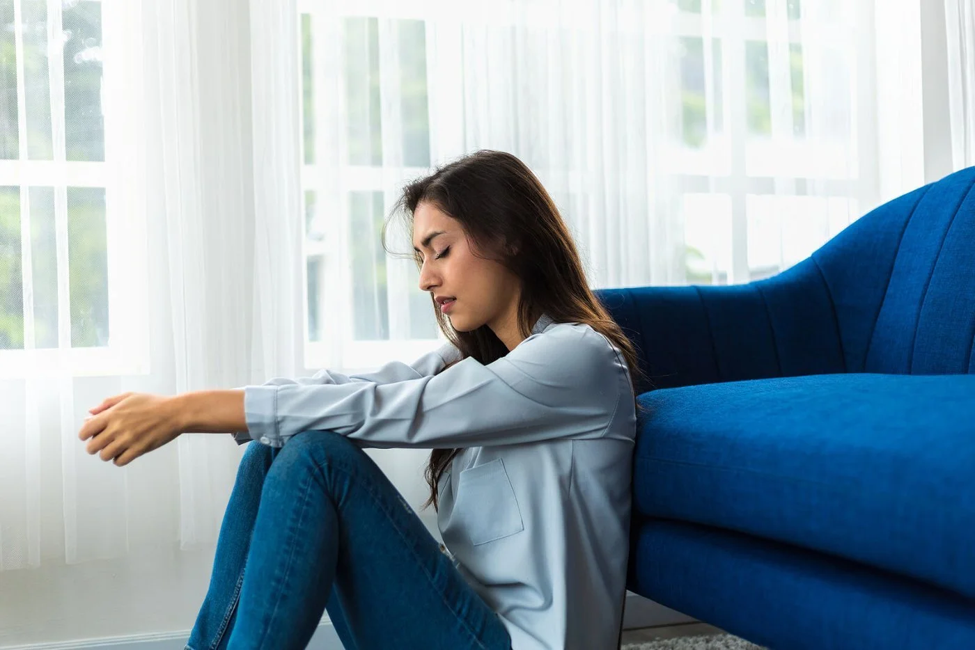 A woman sits on the floor leaning against a blue sofa beside sheer curtains, eyes closed and arms resting on her knees in a quiet moment of grief