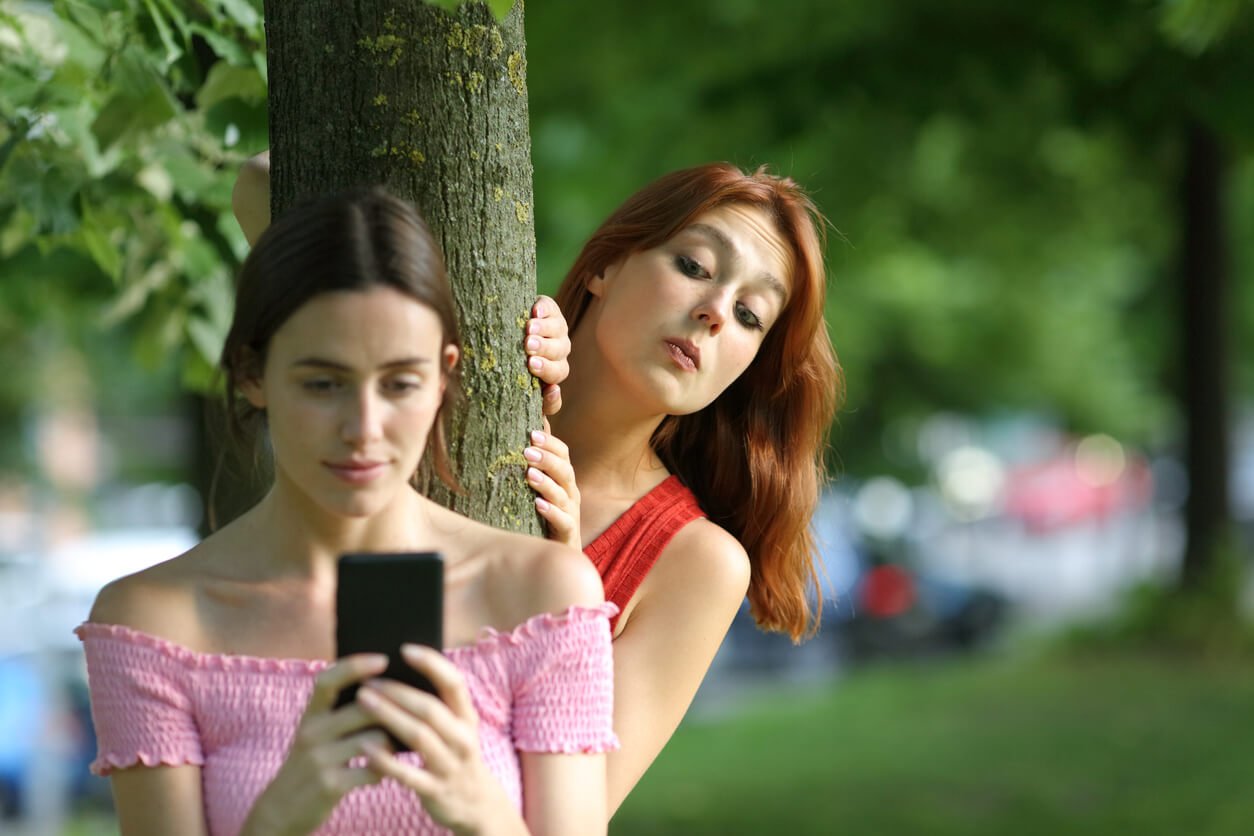 A woman looks at her phone beside a tree while another woman peeks out from behind her shoulder, showing how easily startled highly sensitive people can be.